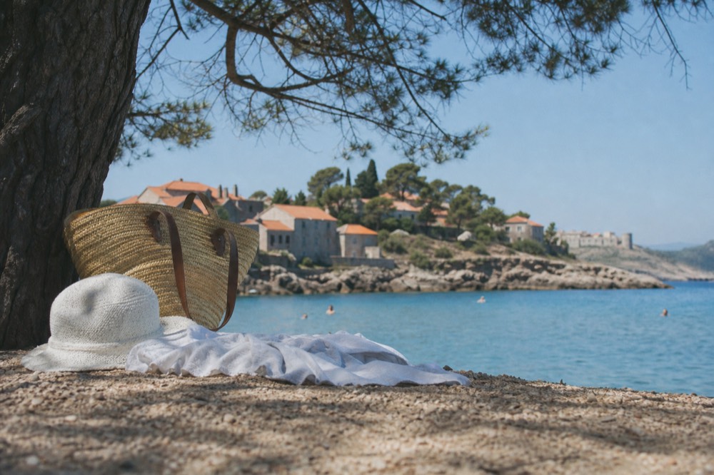 A beach bag, hat and towel on sand under a pine tree
