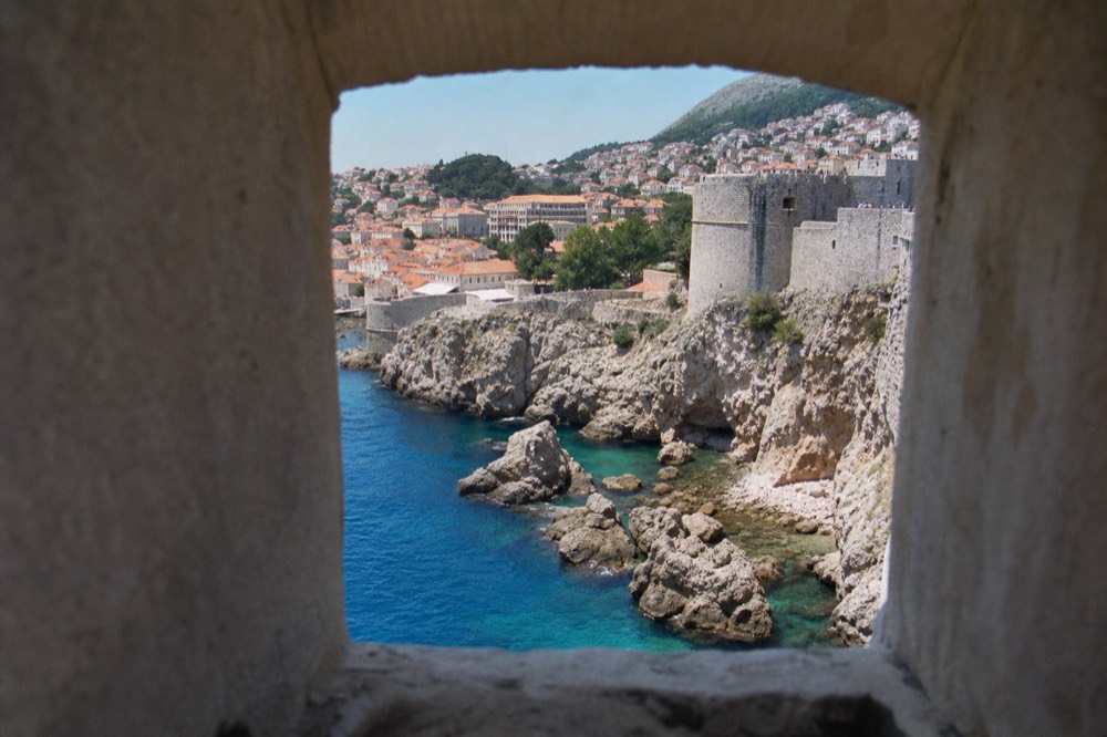 Rocky shoreline with clear turquoise water and a walled town in the distance