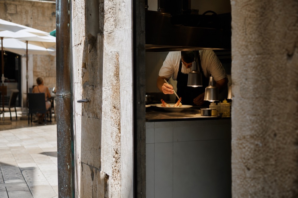 Chef plating a seafood dish with tweezers in a restaurant kitchen pass