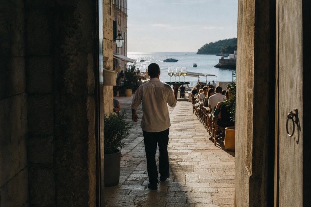 Waiter carrying a tray of wine glasses through a narrow stone alley restaurant terrace
