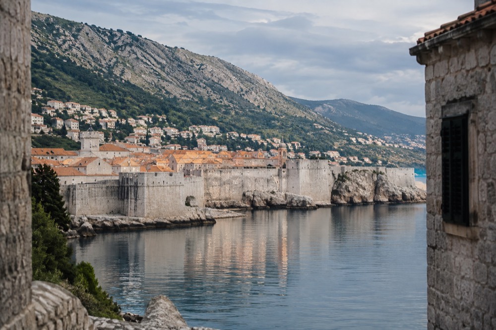 A walled coastal town at the foot of steep mountains reflected in a calm bay