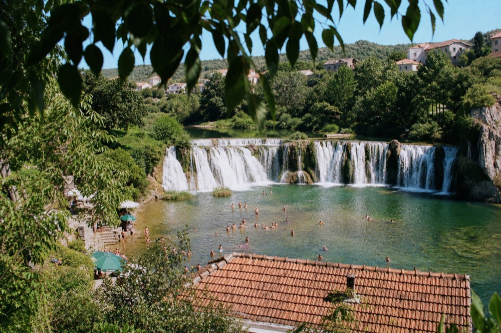 People swimming in a pool below a wide rocky waterfall surrounded by green trees