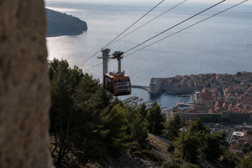A cable car cabin travelling up the steep cableway above pine trees toward the upper station on Mount Srd