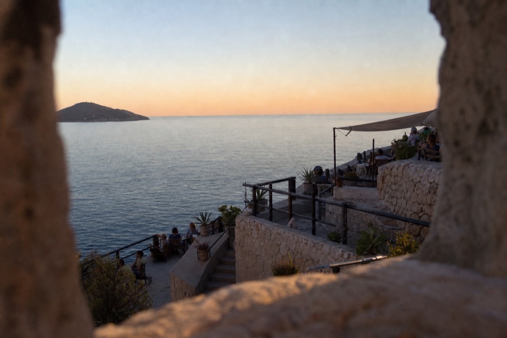 A cliffside bar terrace above the sea at dusk with the island silhouette on the horizon