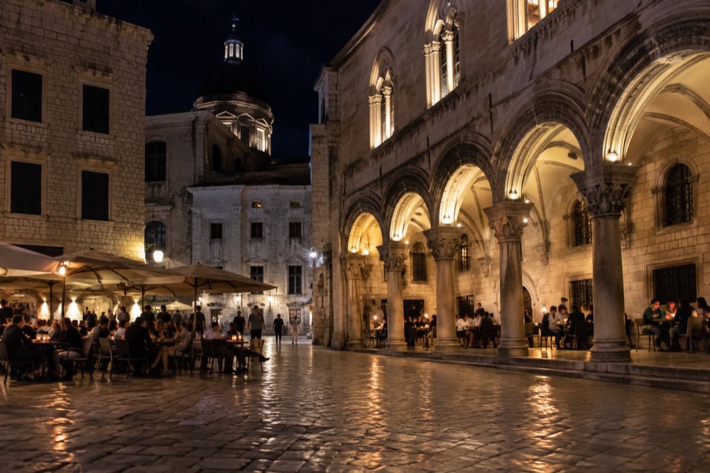 A stone square at night with lit arches and people sitting at bar tables