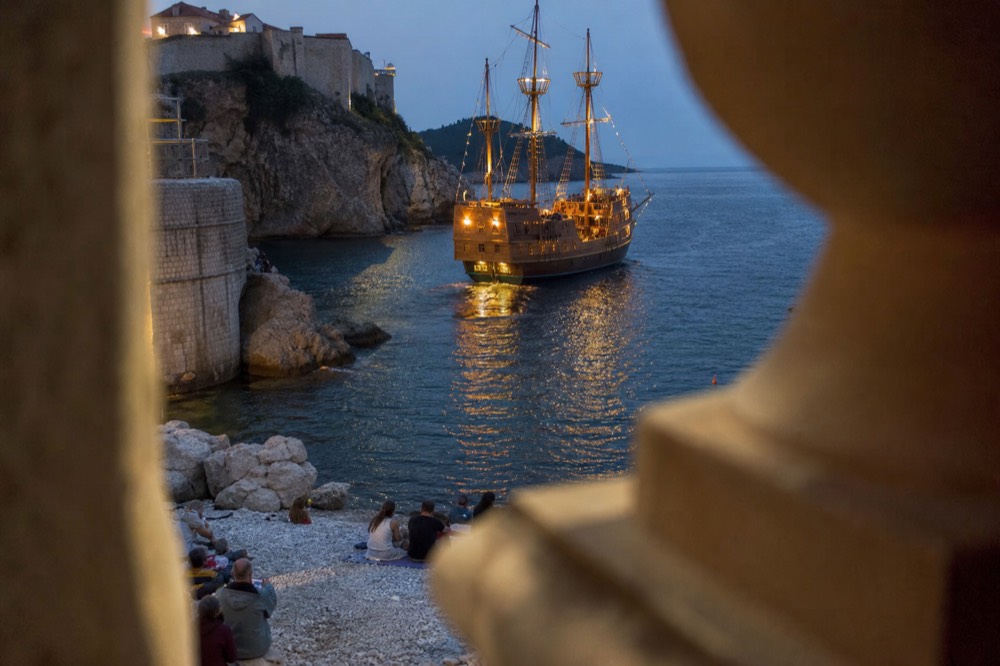 A wooden sailing ship lit up on dark water near the Dubrovnik city walls at dusk