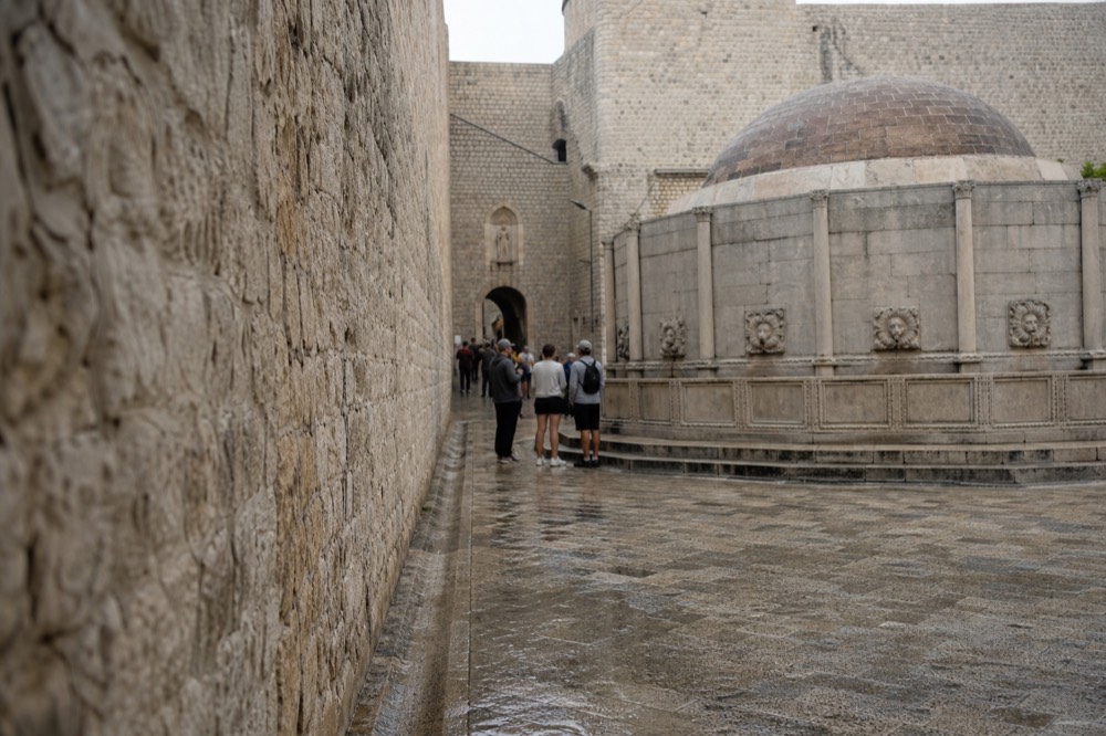 Small groups of visitors standing beside a large stone fountain at the entrance to an old walled city
