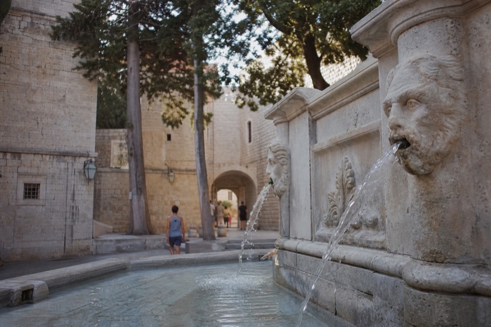 Clear water streaming from a carved stone mask into a wide fountain pool in an old city square