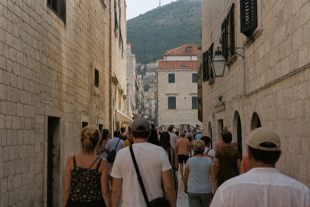 A crowded old town street filled with visitors walking between stone buildings in bright daylight