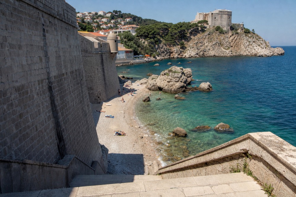 A pebble beach with clear turquoise water below old stone city walls in bright sunlight