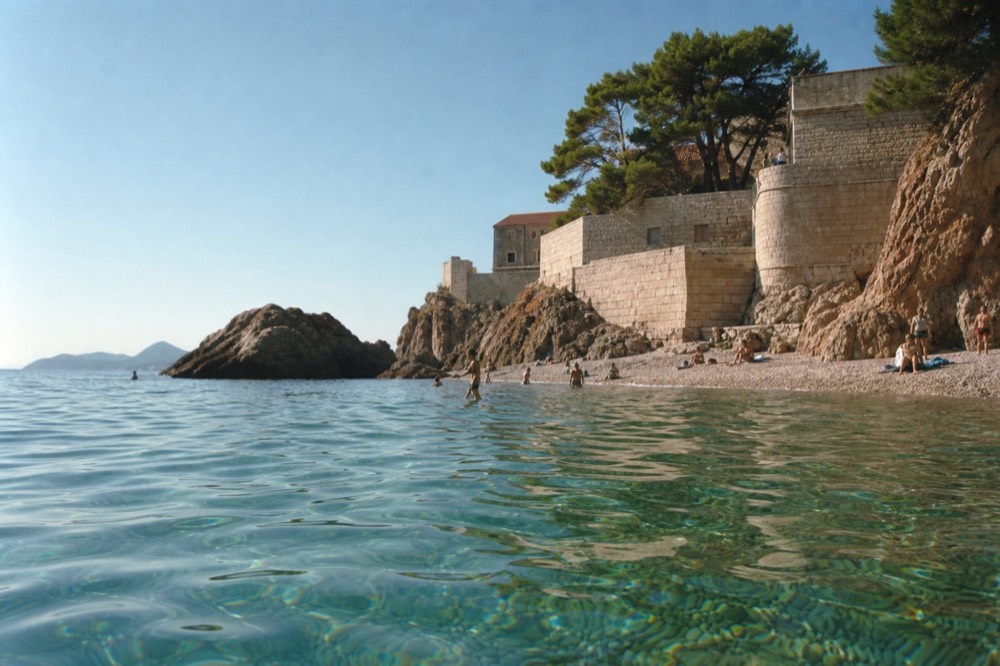 A pebble beach with clear turquoise water and swimmers near stone cliffs