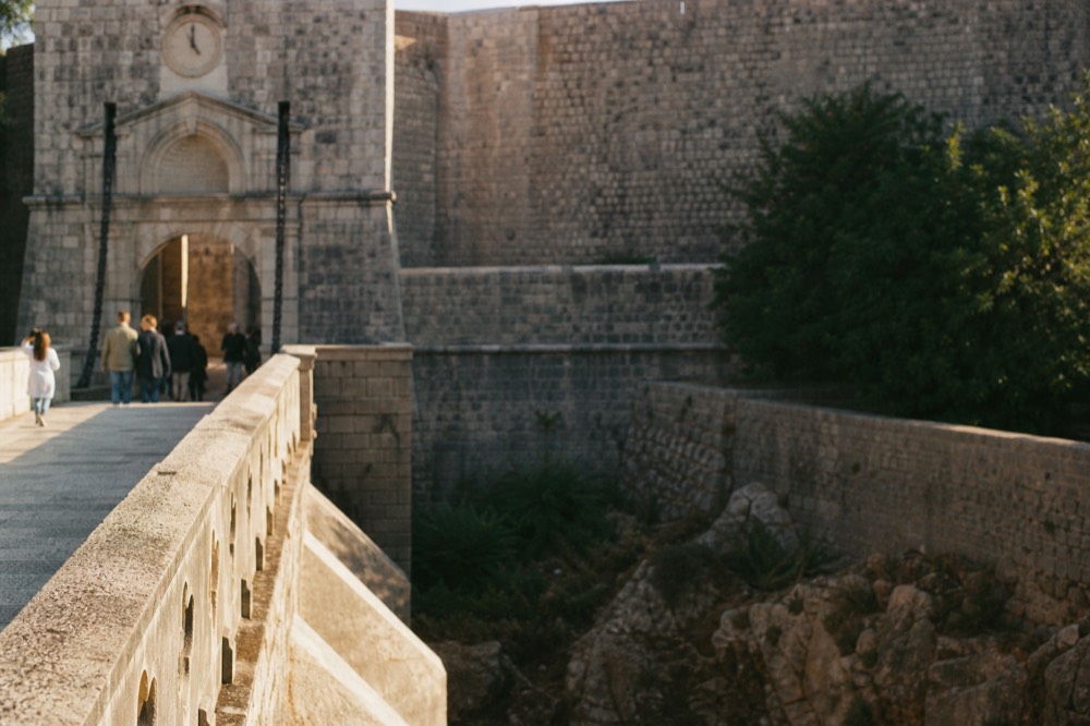 Outer stone bridge crossing a dry moat toward a Renaissance gate with an old clock above it