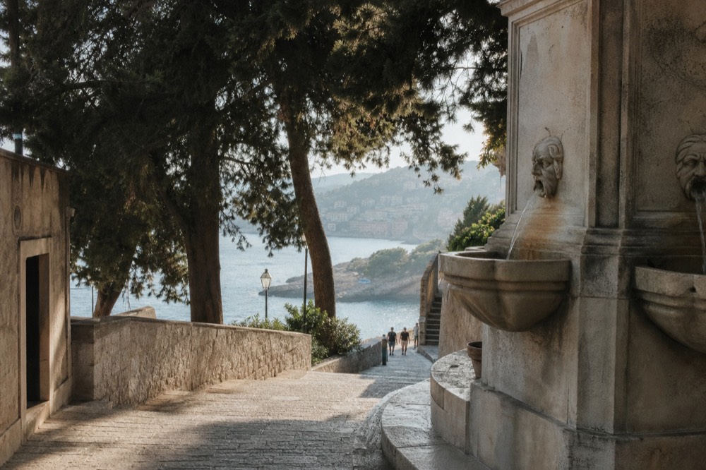 Large stone fountain with carved mask spouts beside a wide limestone street