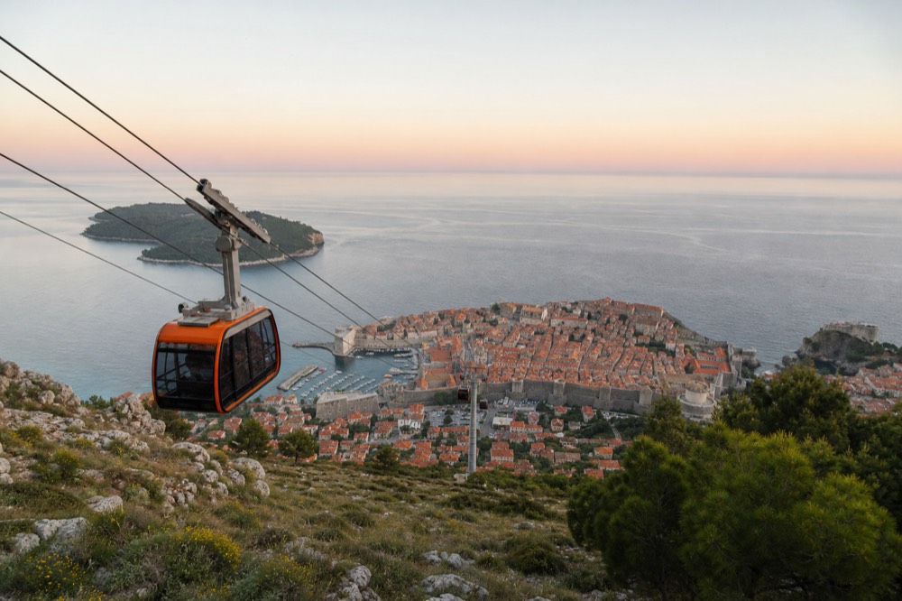 Cable car cabins ascending a steep hillside above a walled coastal town at sunset