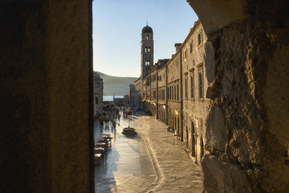Wide limestone-paved street with baroque facades and a bell tower at the far end