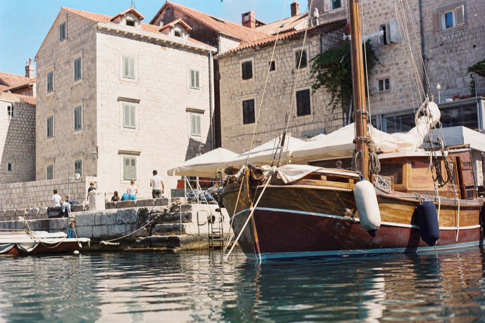 A wooden sailboat moored in a small Adriatic harbour with stone buildings behind it
