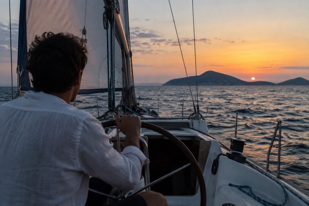 A person at the helm of a small sailboat with islands in the distance