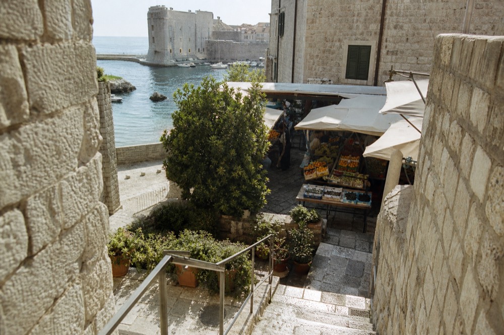 An outdoor market square with crates of figs, oranges and herbs under awnings