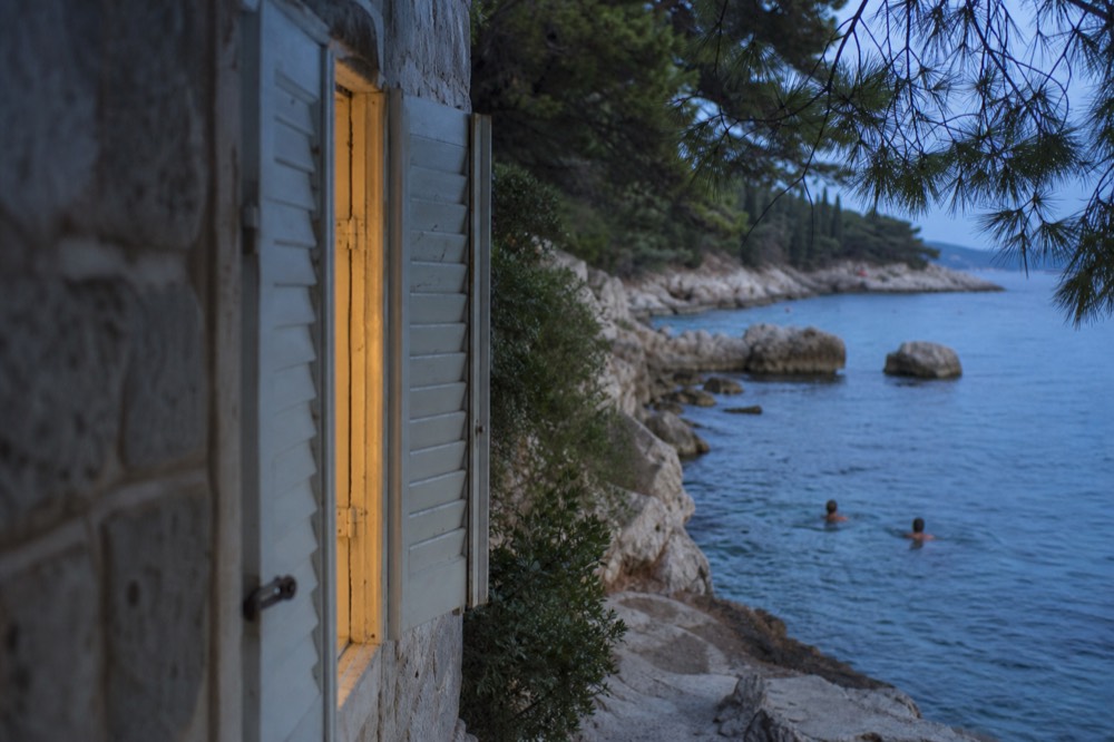 Swimmers in clear blue water beside smooth rocks under pine trees