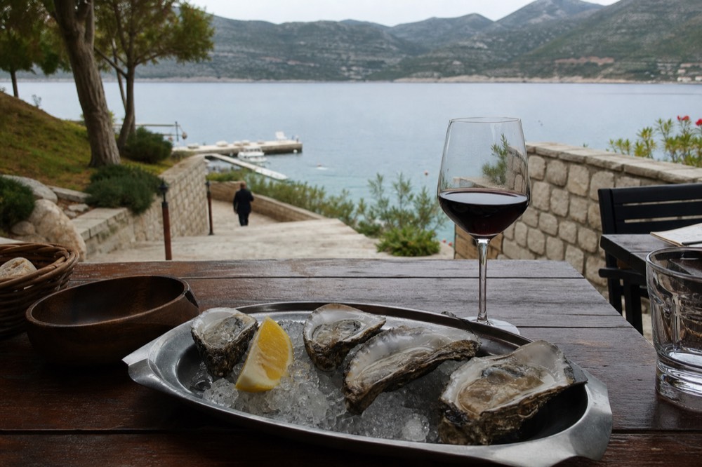 Oysters opened on a tray beside a glass of red wine at a waterfront table