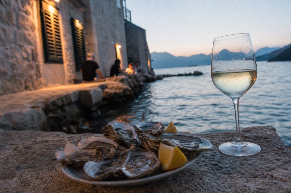 Fresh oysters on a plate beside a glass of white wine at a waterside table