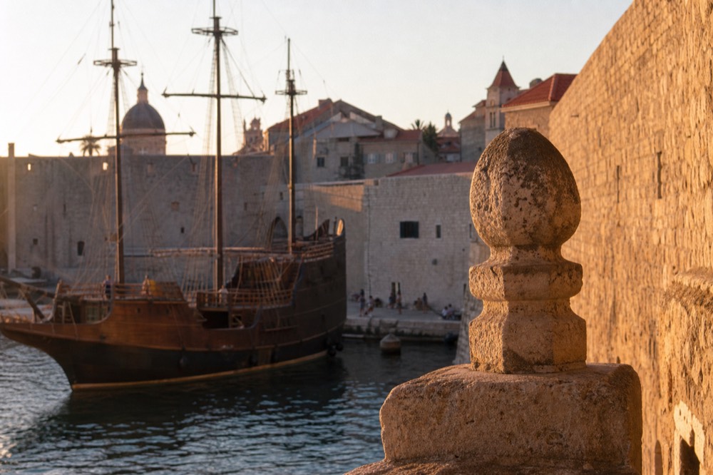 A wooden sailing ship with masts moored in Dubrovnik's old harbour at golden hour