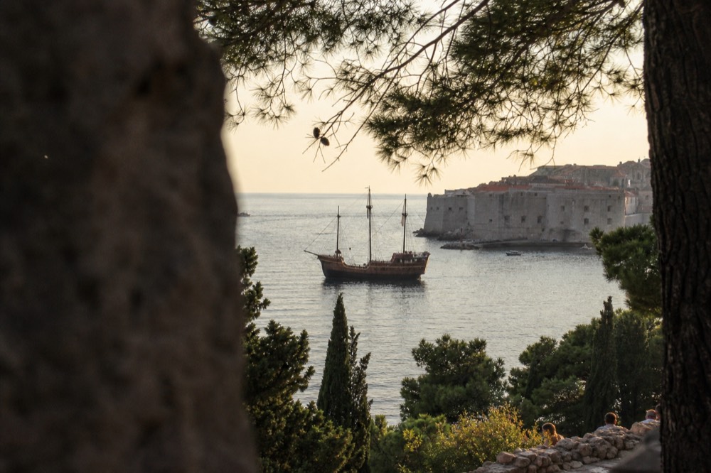 A wooden sailing ship with tall masts on calm water near a walled coastal city at dusk
