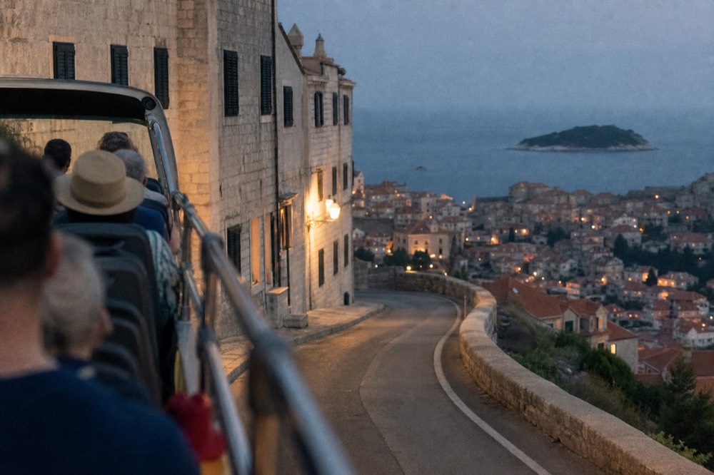 An open-top bus on a coastal road above a red-roofed town at dusk