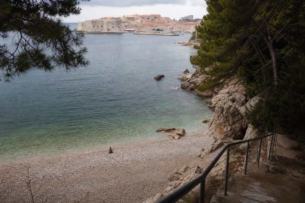 A quiet pebble cove with clear shallow water and a view of Dubrovnik's walled Old Town across the bay
