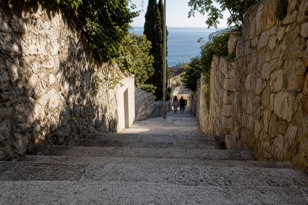 A shaded residential lane with stone walls leading downhill toward the sea