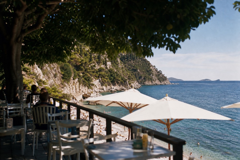 A cafe terrace with tables and umbrellas overlooking a pebble beach and the sea