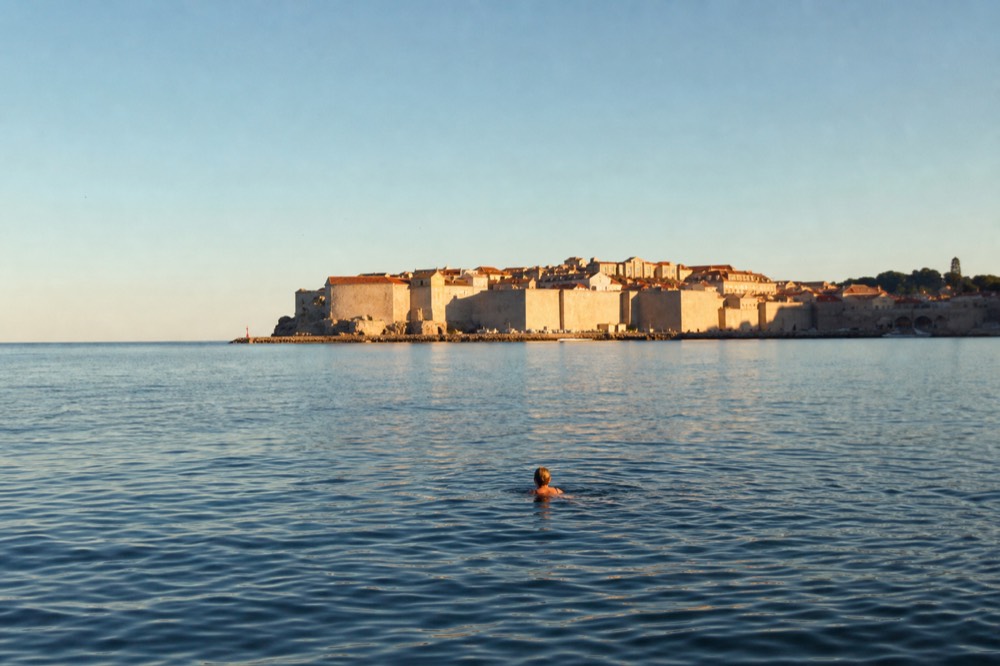 Swimmer floating on calm evening sea with the walled city glowing in background light