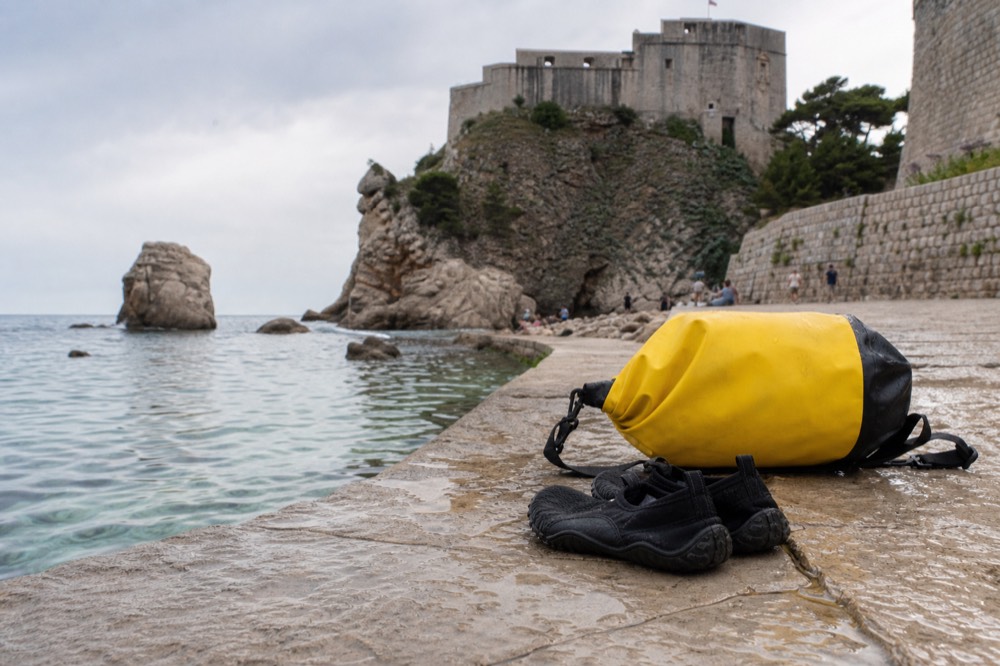 Dry bag and water shoes on a stone ledge beside clear sea water