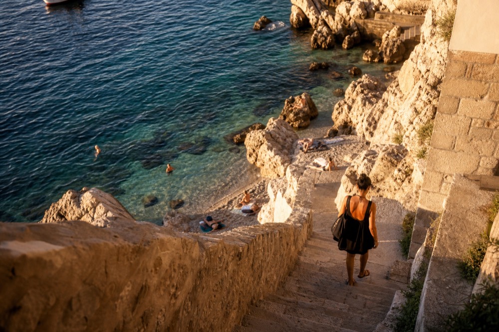 The rocky shoreline of Sveti Jakov beach with clear turquoise water and a few sunbathers.