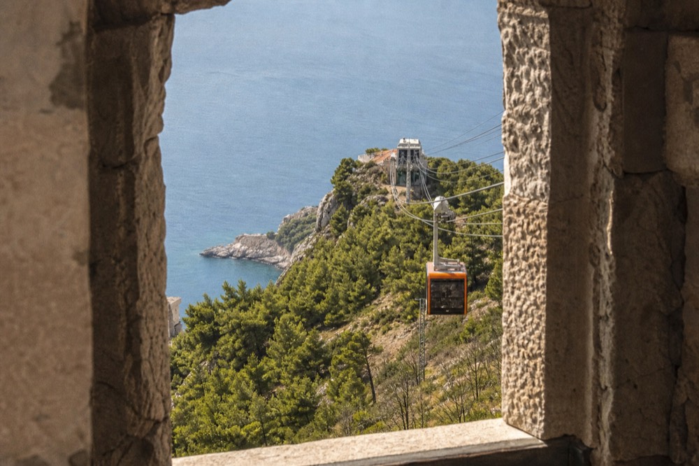 A cable car cabin ascending above pine slopes toward a hilltop station above the sea