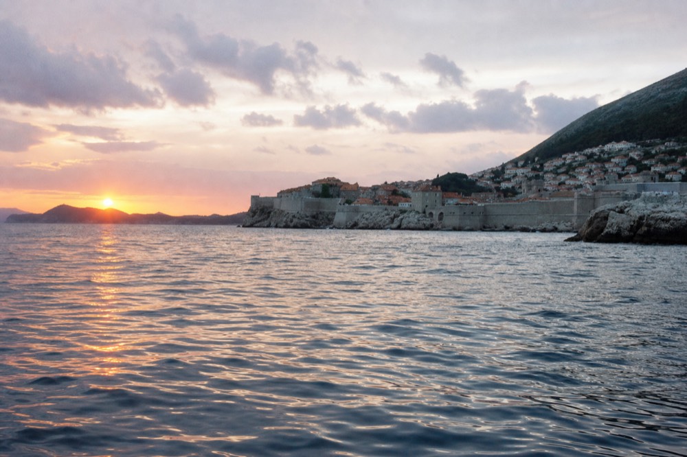 The sun setting over islands behind a walled coastal city seen from the sea