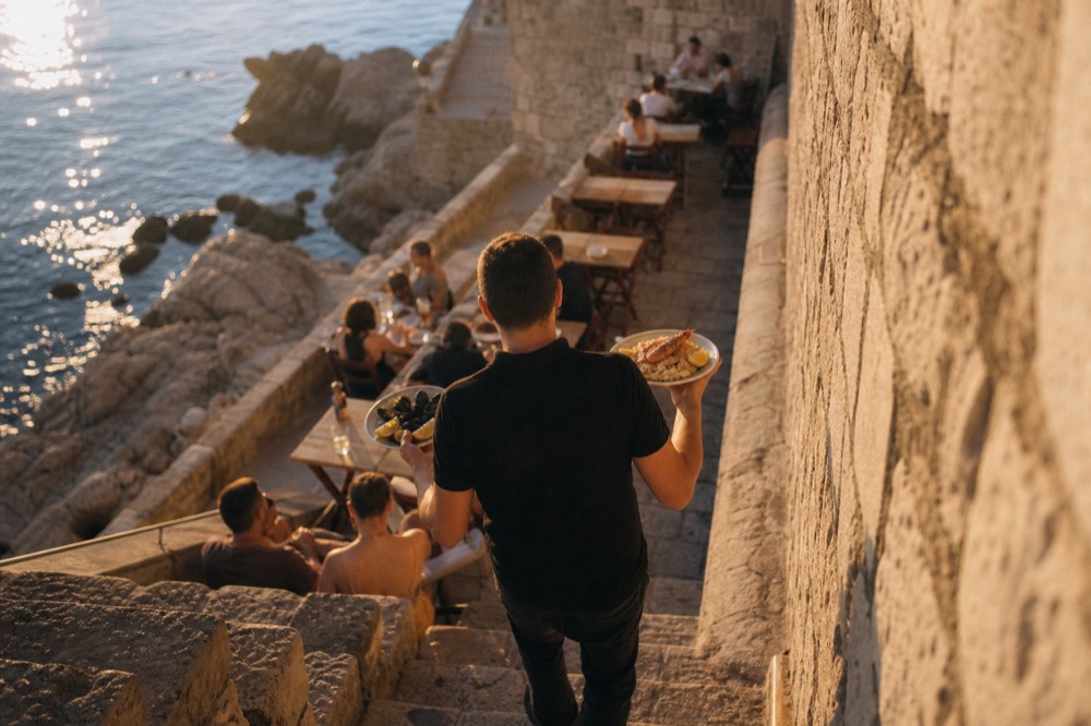 A waiter carrying plates of seafood to a table on a stone terrace at dusk